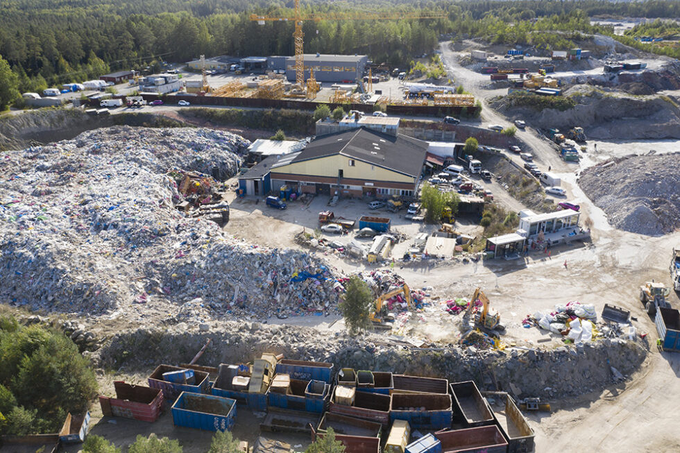 Drönarbild som visar en anläggning med byggnader, containrar och stora högar med byggavfall.