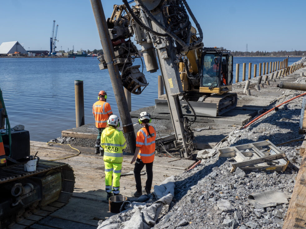 Tre personer i varselkläder och hjälmar som tittar på pålningsmaskinen som arbetar på kajen vid anläggandet av Skellefteå Port.