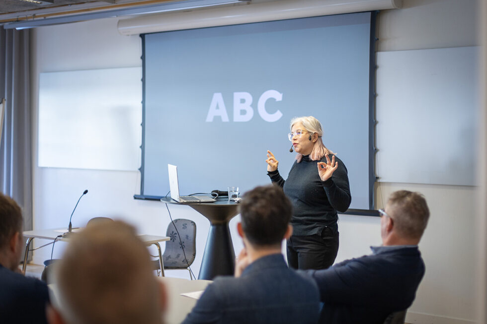 kvinna som står framför en skärm eller duk som det står ABC på. Man ser också ryggarna på några personer.