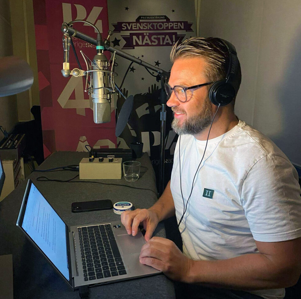 Kim Söderström sitter i radiostudion och spelar in sitt sommarprat. En mikrofon hänger ned, en laptop på skrivbordet och i bakgrunden syns skyltar som gör reklam för Svensktoppen nästa.